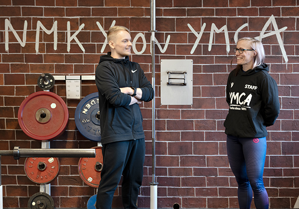 Kaksi ihmistä seisovat seinän edessä, taustalla NMKY on YMCA -teksti