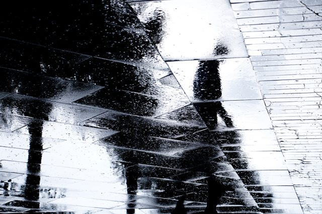 Blurry reflection silhouette of one person walking alone on wet city park sidewalk on a rainy day